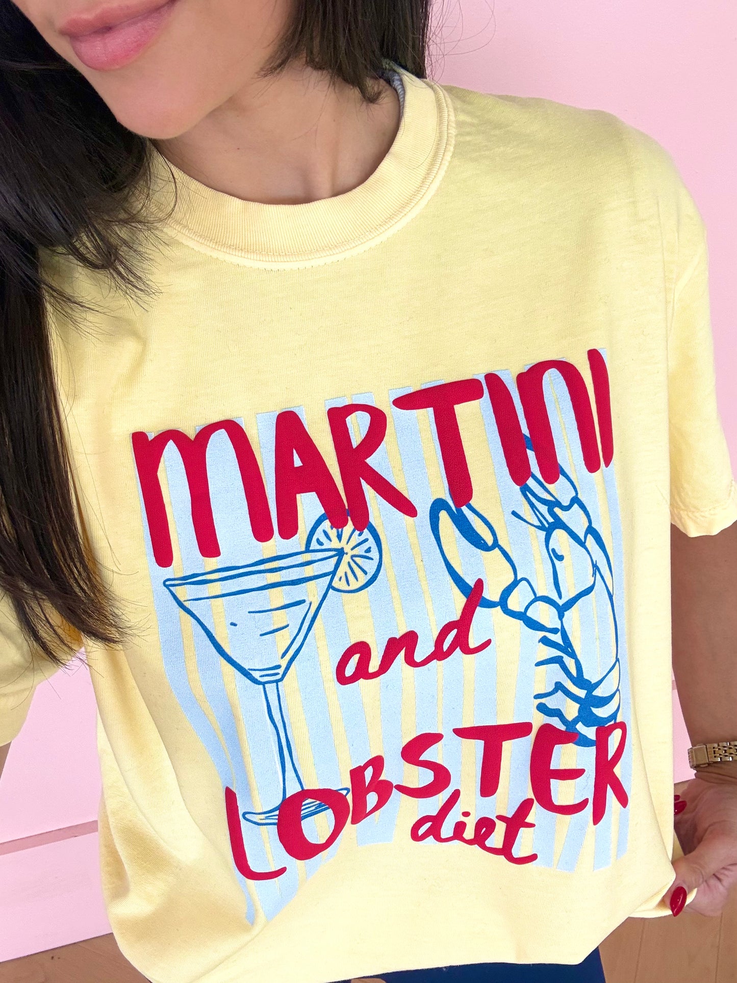 A close-up of a person wearing a pale yellow graphic T-shirt with the words 'MARTINI and LOBSTER diet' in red, plus a martini glass and lobster illustration in blue, against a pink background.