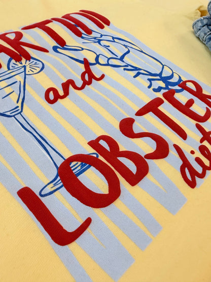 A close-up of a yellow knit garment featuring a colorful graphic print with red text reading 'MARTINI and LOBSTER' and blue striped accents.