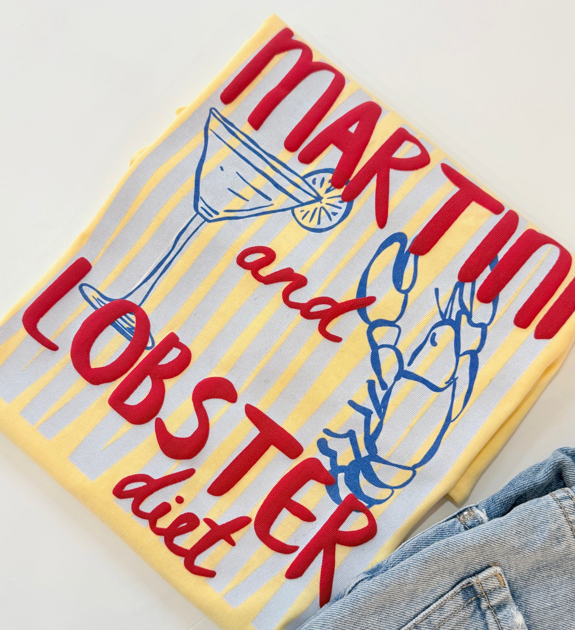 A folded graphic T-shirt with a yellow and white striped background, featuring the text 'MARTINI and LOBSTER diet' in red script, a martini glass illustration, and a blue lobster drawing. A pair of blue jeans is partially visible at the bottom right.
