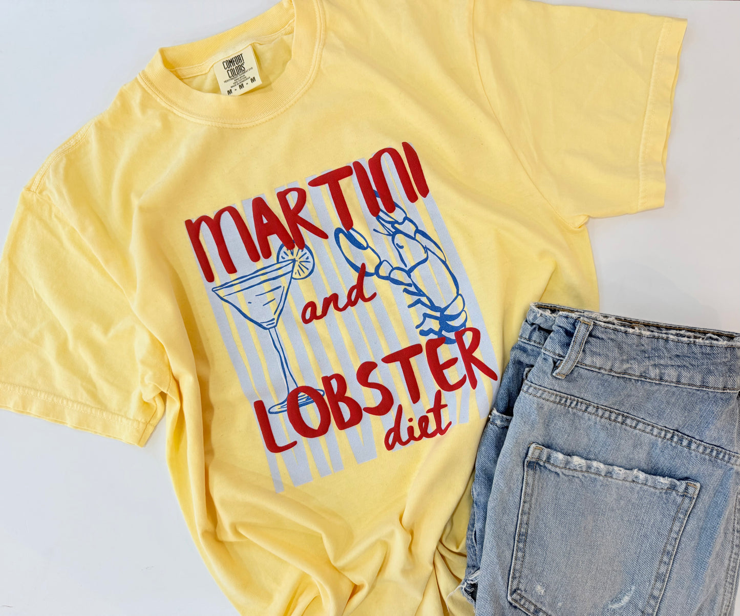 A yellow graphic t-shirt featuring the text 'MARTINI and LOBSTER diet' in red and blue, styled flat-lay next to light-wash blue jeans on a white background.