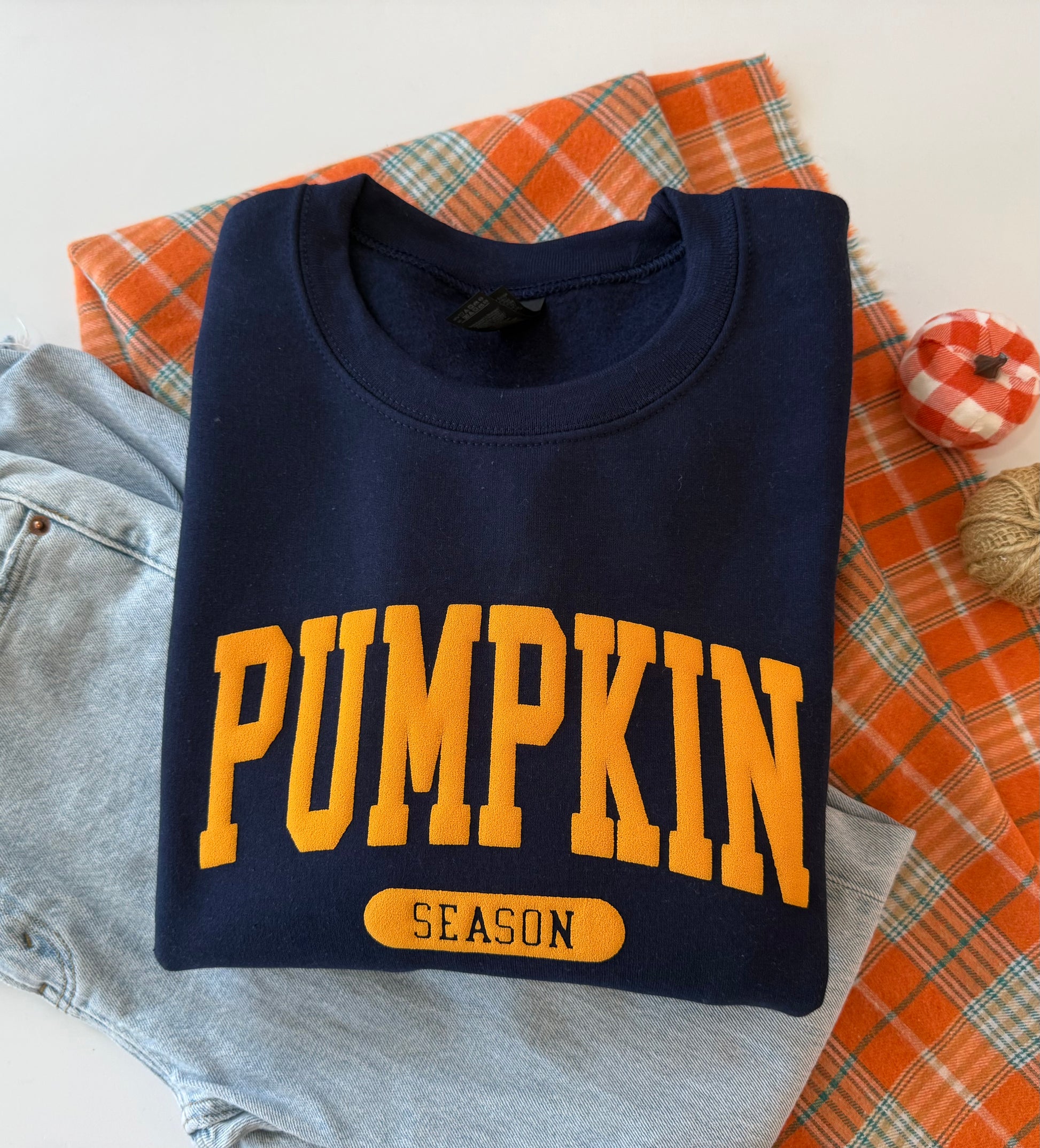 A folded navy blue sweatshirt with 'PUMPKIN SEASON' printed in yellow on the front, styled on top of light blue jeans, an orange plaid fabric, and a small decorative pumpkin.