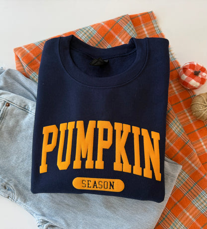 A folded navy blue sweatshirt with 'PUMPKIN SEASON' printed in yellow on the front, styled on top of light blue jeans, an orange plaid fabric, and a small decorative pumpkin.
