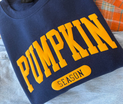 A navy blue crewneck sweatshirt with 'PUMPKIN SEASON' printed in yellow varsity-style lettering, laid flat on a light gray surface.