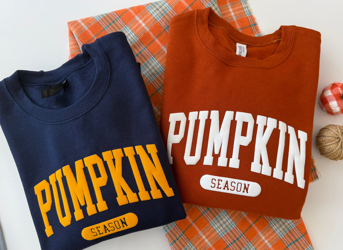 Two folded crewneck sweatshirts displayed on an orange plaid fabric background. The left sweatshirt is navy blue with yellow 'PUMPKIN SEASON' text, and the right sweatshirt is rust orange with white 'PUMPKIN SEASON' text.