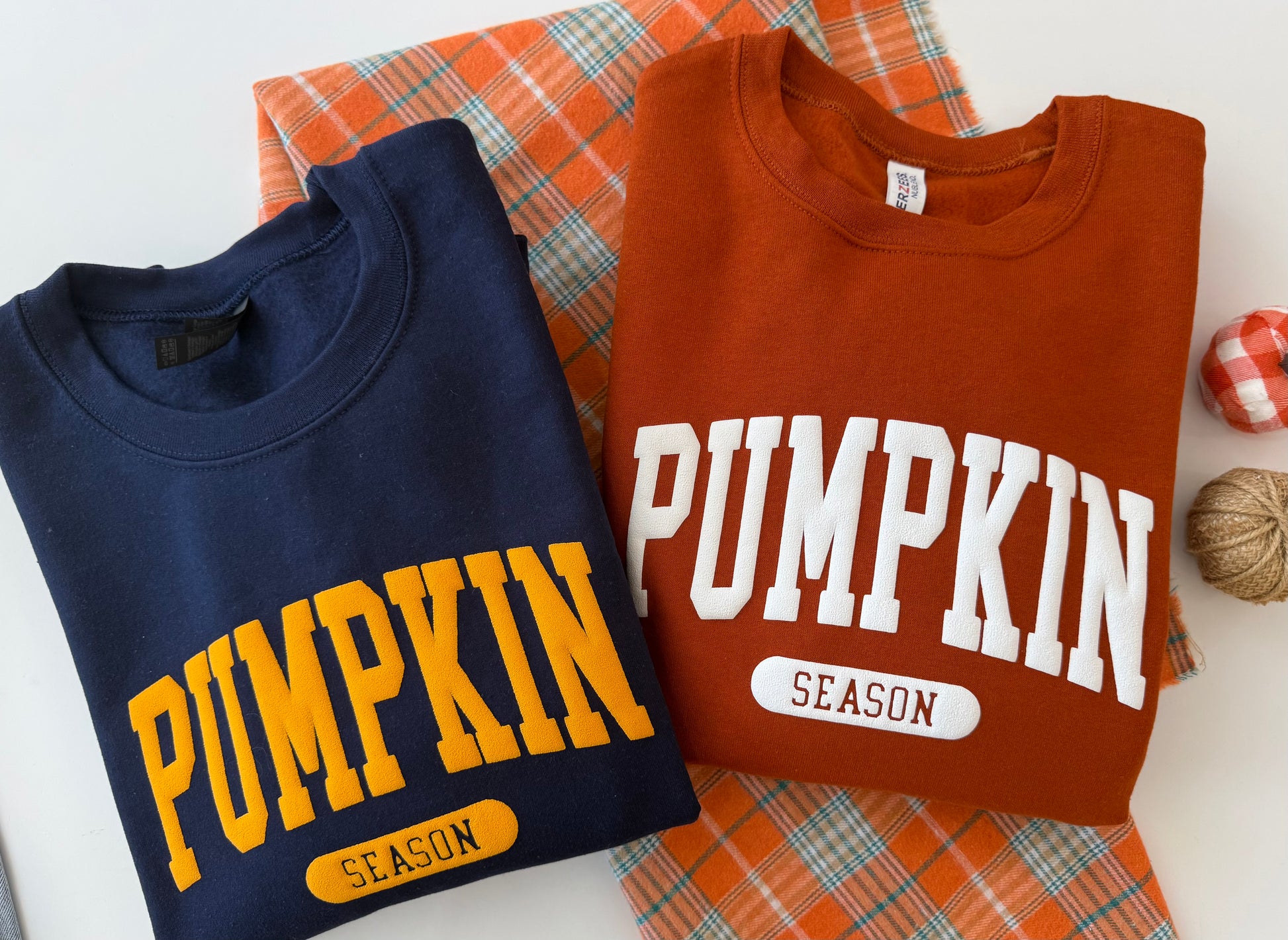Two folded crewneck sweatshirts displayed on an orange plaid fabric background. The left sweatshirt is navy blue with yellow 'PUMPKIN SEASON' text, and the right sweatshirt is rust orange with white 'PUMPKIN SEASON' text.