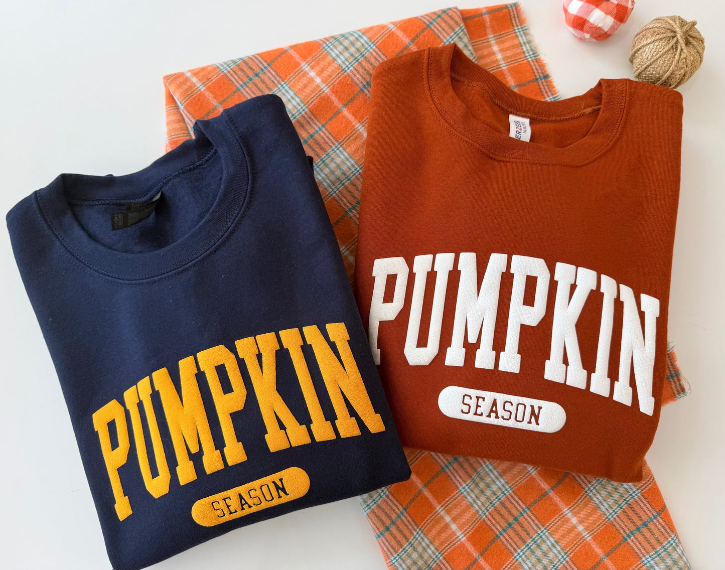 Two folded crewneck sweatshirts displayed on a white background with an orange plaid fabric and autumn props. The left sweatshirt is navy blue with yellow 'PUMPKIN SEASON' lettering, and the right sweatshirt is rust orange with white 'PUMPKIN SEASON' lettering.