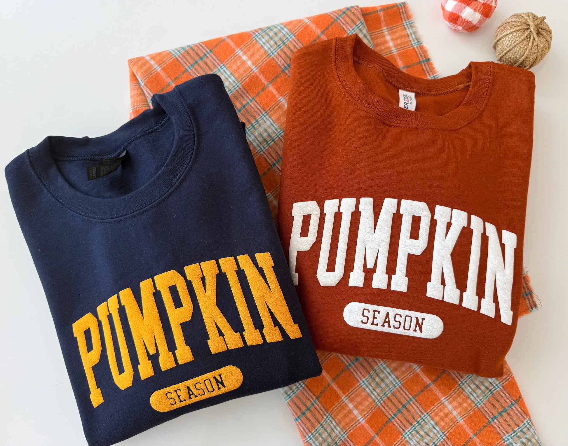 Two folded crewneck sweatshirts displayed on a white background with an orange plaid fabric and autumn props. The left sweatshirt is navy blue with yellow 'PUMPKIN SEASON' lettering, and the right sweatshirt is rust orange with white 'PUMPKIN SEASON' lettering.