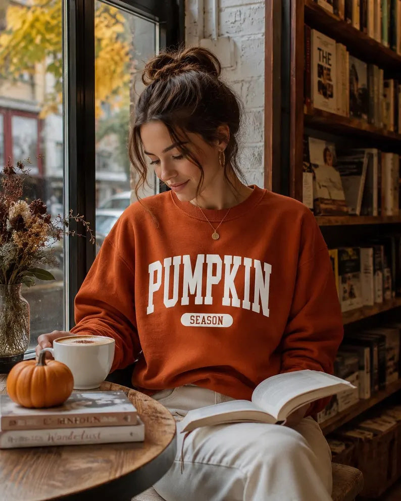 A woman sitting in a cozy bookstore or cafe setting, wearing a rust-orange crewneck sweatshirt with 'PUMPKIN SEASON' printed in white on the front. She is holding an open book and looking down, with a small pumpkin, a mug, and bookshelves visible in the background.
