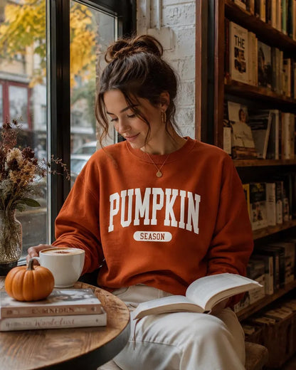 A woman sitting in a cozy bookstore or cafe setting, wearing a rust-orange crewneck sweatshirt with 'PUMPKIN SEASON' printed in white on the front. She is holding an open book and looking down, with a small pumpkin, a mug, and bookshelves visible in the background.