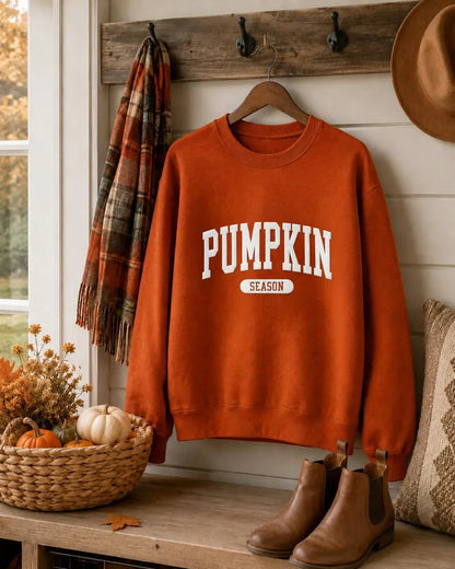 An orange crewneck sweatshirt with 'PUMPKIN SEASON' printed in white on the front, hanging on a wooden hanger on a rustic wall. The scene is styled with a plaid scarf, a basket of pumpkins, brown boots, and a hat.