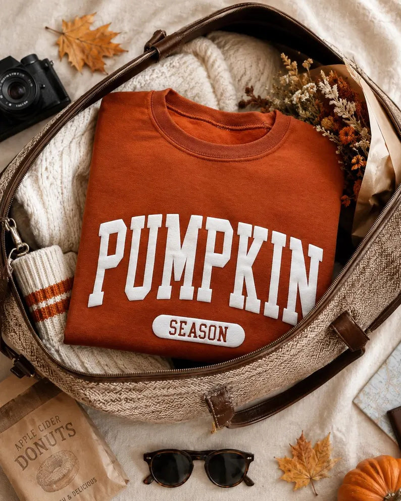 An orange crewneck sweatshirt with 'PUMPKIN SEASON' printed in white varsity lettering, neatly folded inside a beige woven suitcase alongside a cream knit sweater, sunglasses, a camera, a small pumpkin, and autumn leaves.