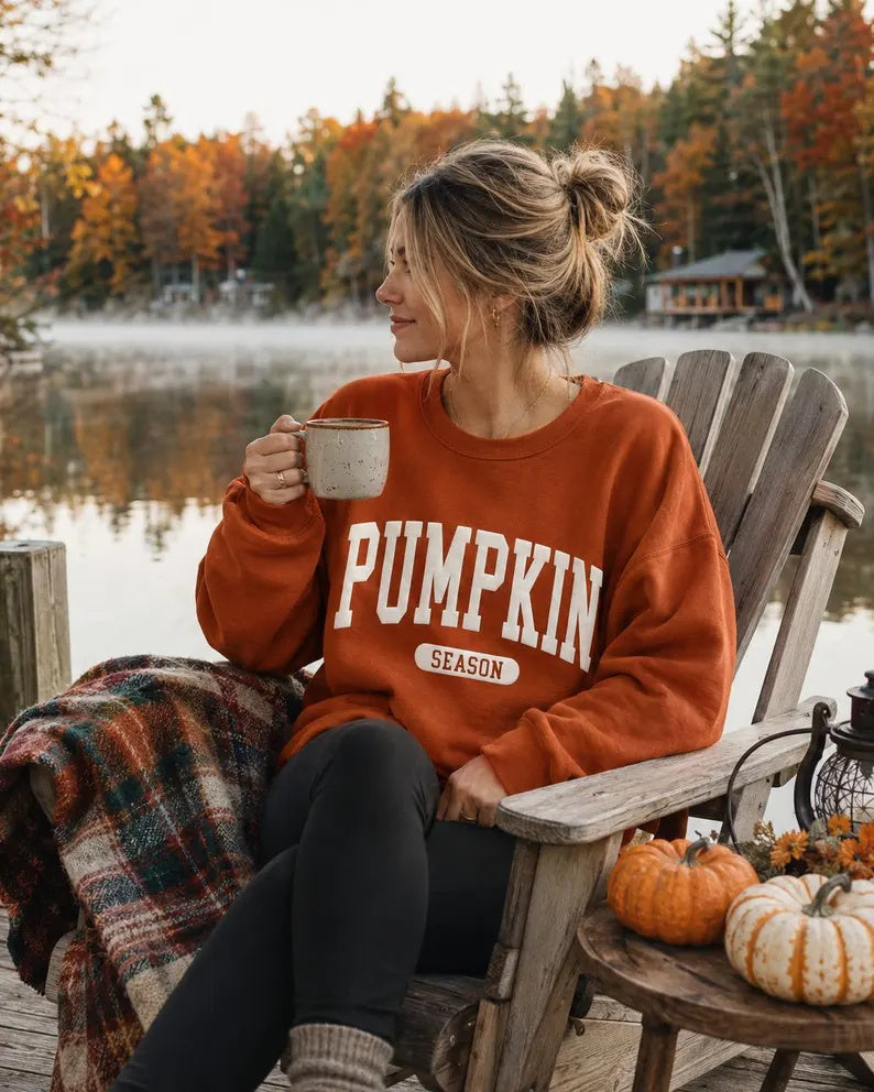 A woman sitting in a wooden Adirondack chair by a lake during autumn, wearing an orange crewneck sweatshirt with 'PUMPKIN SEASON' printed in white. She holds a mug, with pumpkins on a side table and a plaid blanket draped over her lap.