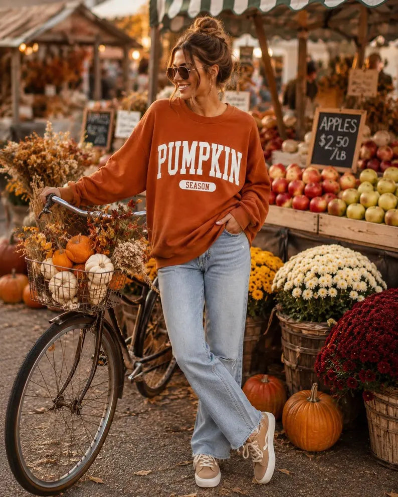 A woman stands at an outdoor autumn market wearing a rust-orange crewneck sweatshirt with 'PUMPKIN SEASON' printed in white across the front, paired with light blue jeans and beige sneakers. She is leaning against a bicycle filled with pumpkins, with apple stands and flowers in the background.