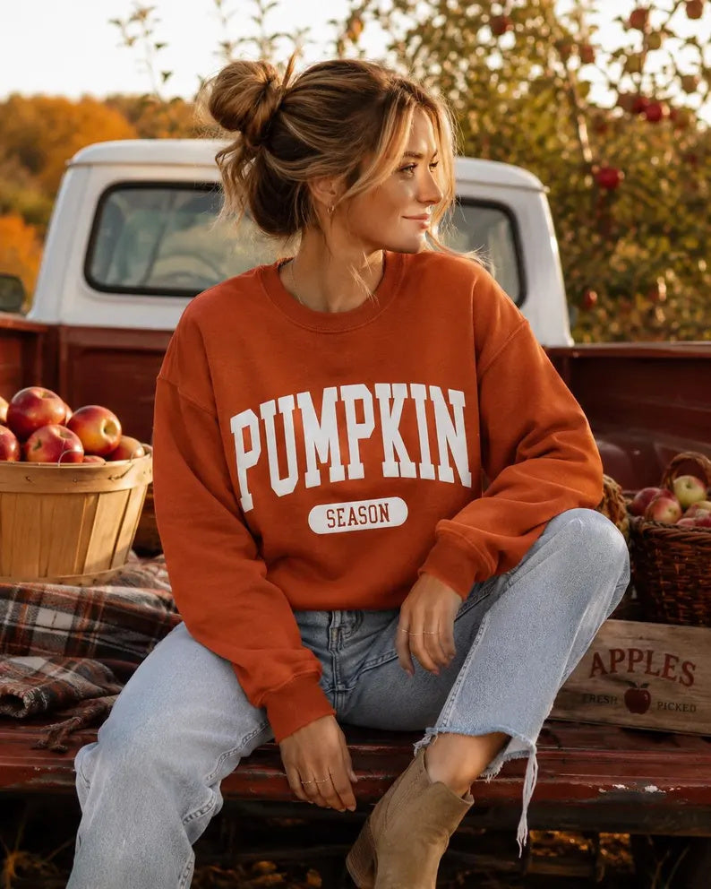 A woman sits in the bed of a pickup truck surrounded by apples, wearing a rust-orange crewneck sweatshirt with 'PUMPKIN SEASON' printed in white. She is wearing light-wash blue jeans and tan ankle boots.