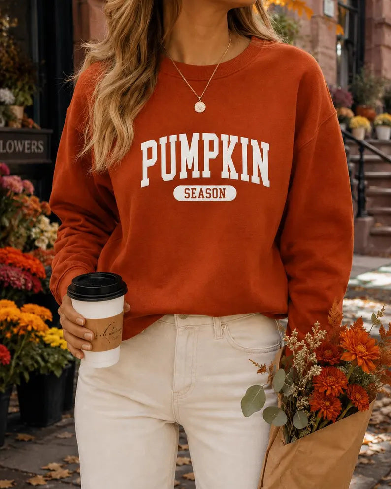 A woman wearing a rust-orange crewneck sweatshirt with 'PUMPKIN SEASON' printed in white on the front, paired with cream pants, holding a coffee cup and a paper bag of flowers in an autumn flower shop setting.