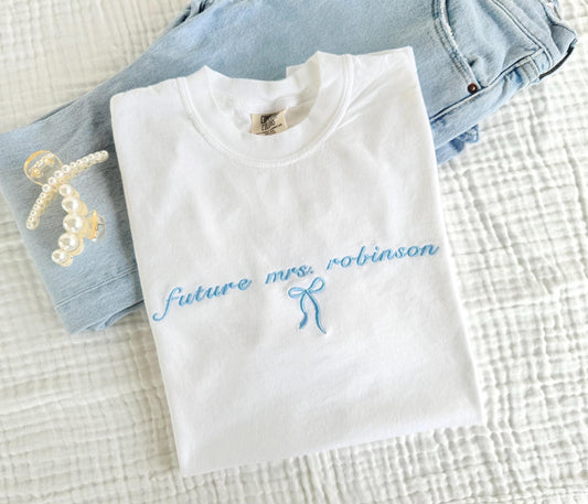 White Comfort Colors t-shirt with light blue embroidered text reading “future mrs. robinson” and a delicate bow, folded on a white textured blanket next to light wash jeans and pearl hair clips – perfect custom bridal shirt or engagement gift.