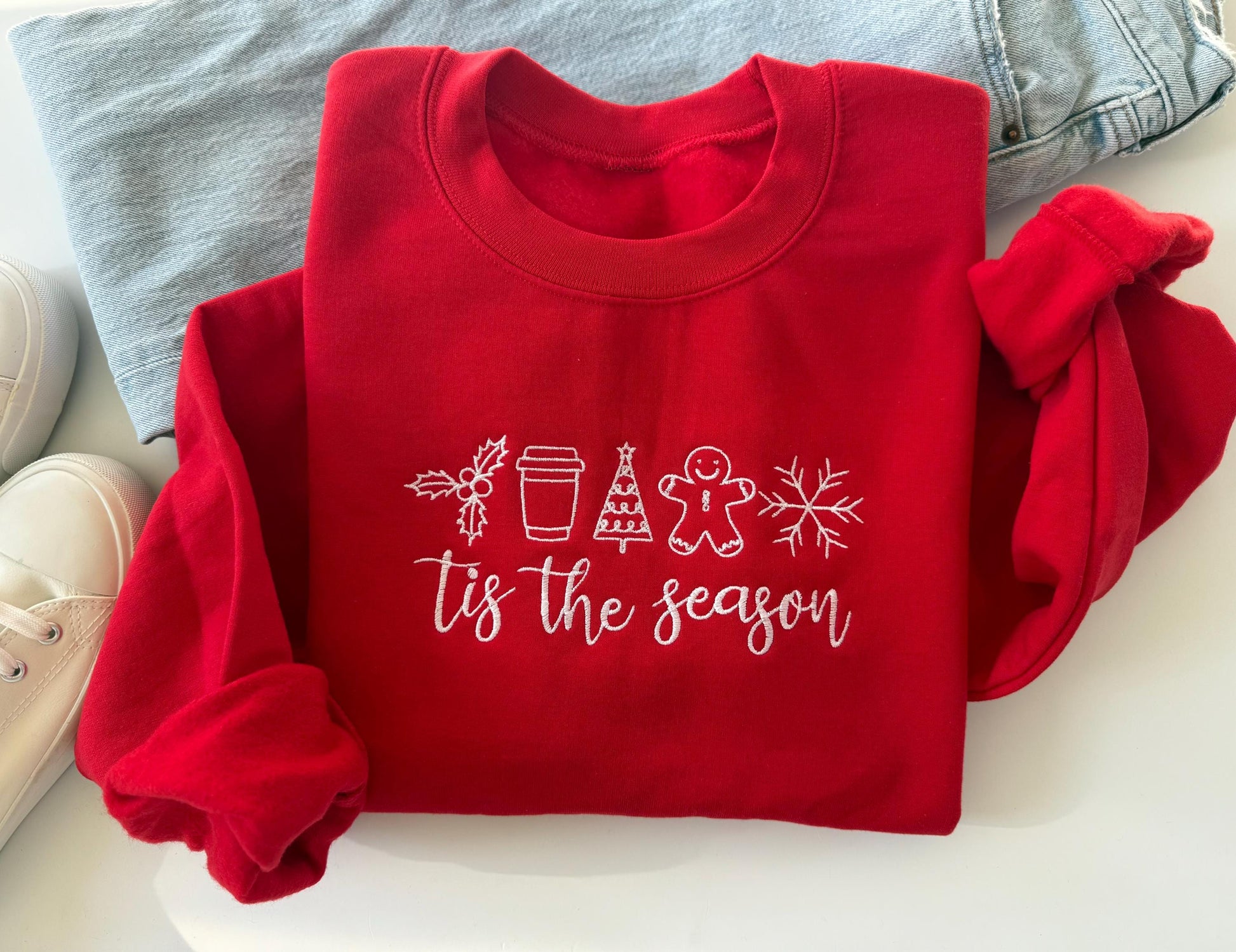 Red embroidered Christmas sweatshirt with “’Tis the Season” text and festive icons including holly, coffee cup, Christmas tree, gingerbread man, and snowflake — cozy holiday crewneck folded on white background with jeans and sneakers.