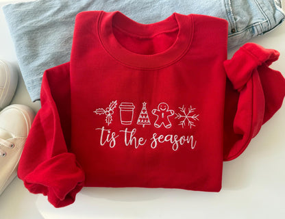 Red embroidered Christmas sweatshirt with “’Tis the Season” text and festive icons including holly, coffee cup, Christmas tree, gingerbread man, and snowflake — cozy holiday crewneck folded on white background with jeans and sneakers.