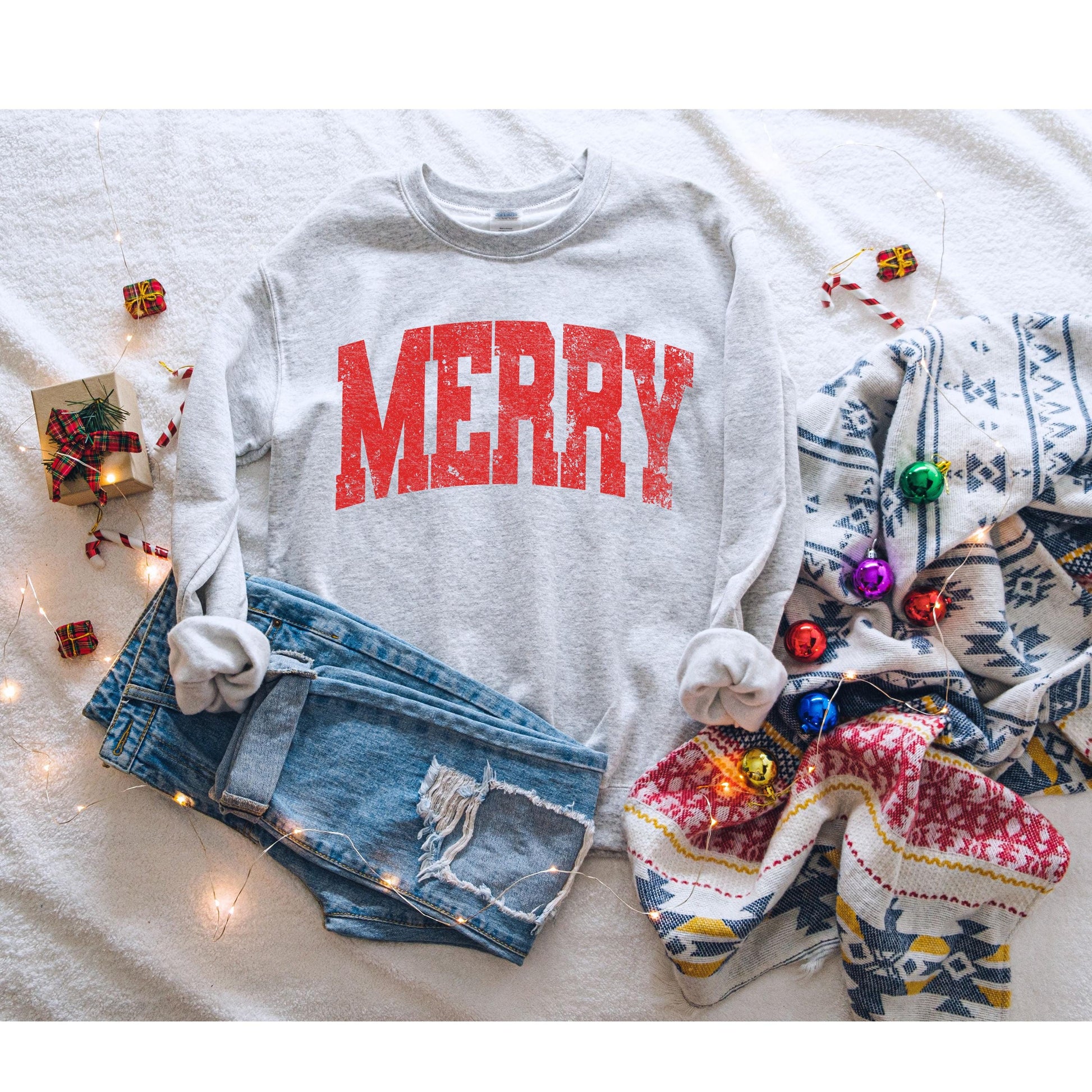 Gray sweatshirt with bold red distressed “MERRY” text on the front, styled next to light-wash jeans and holiday greenery - cozy Christmas crewneck perfect for festive outfits and holiday gifts.