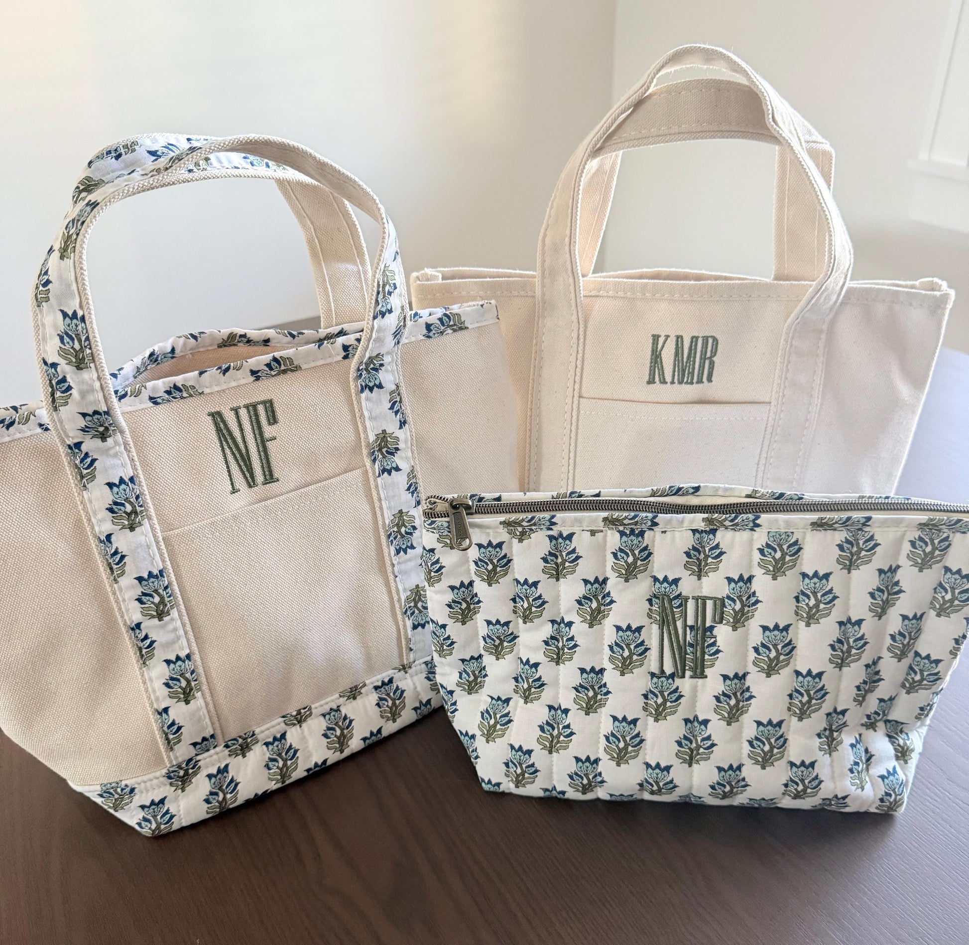 Three canvas items are on a dark wood surface: a small tote with blue floral trim and "NF", a plain tote with "KMR", and a quilted pouch with blue floral pattern and "NF".
