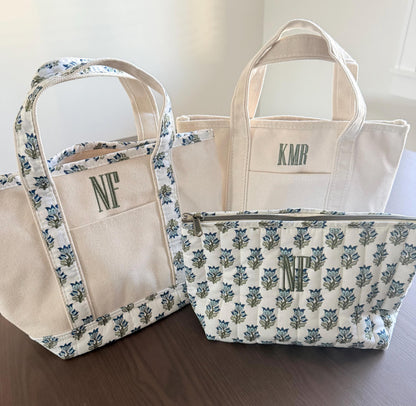 Three canvas items are on a dark wood surface: a small tote with blue floral trim and "NF", a plain tote with "KMR", and a quilted pouch with blue floral pattern and "NF".