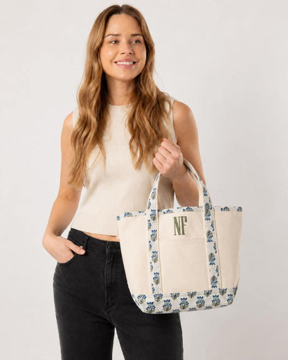 A woman in a cream top and black jeans holds a small canvas tote bag with blue and green floral trim and the green embroidered initials "NF".