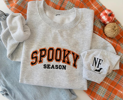 A light gray sweatshirt with "SPOOKY SEASON" embroidered in orange and black, and a cuff with "NF" and a bat, on an orange plaid blanket next to jeans and small decorative pumpkins.