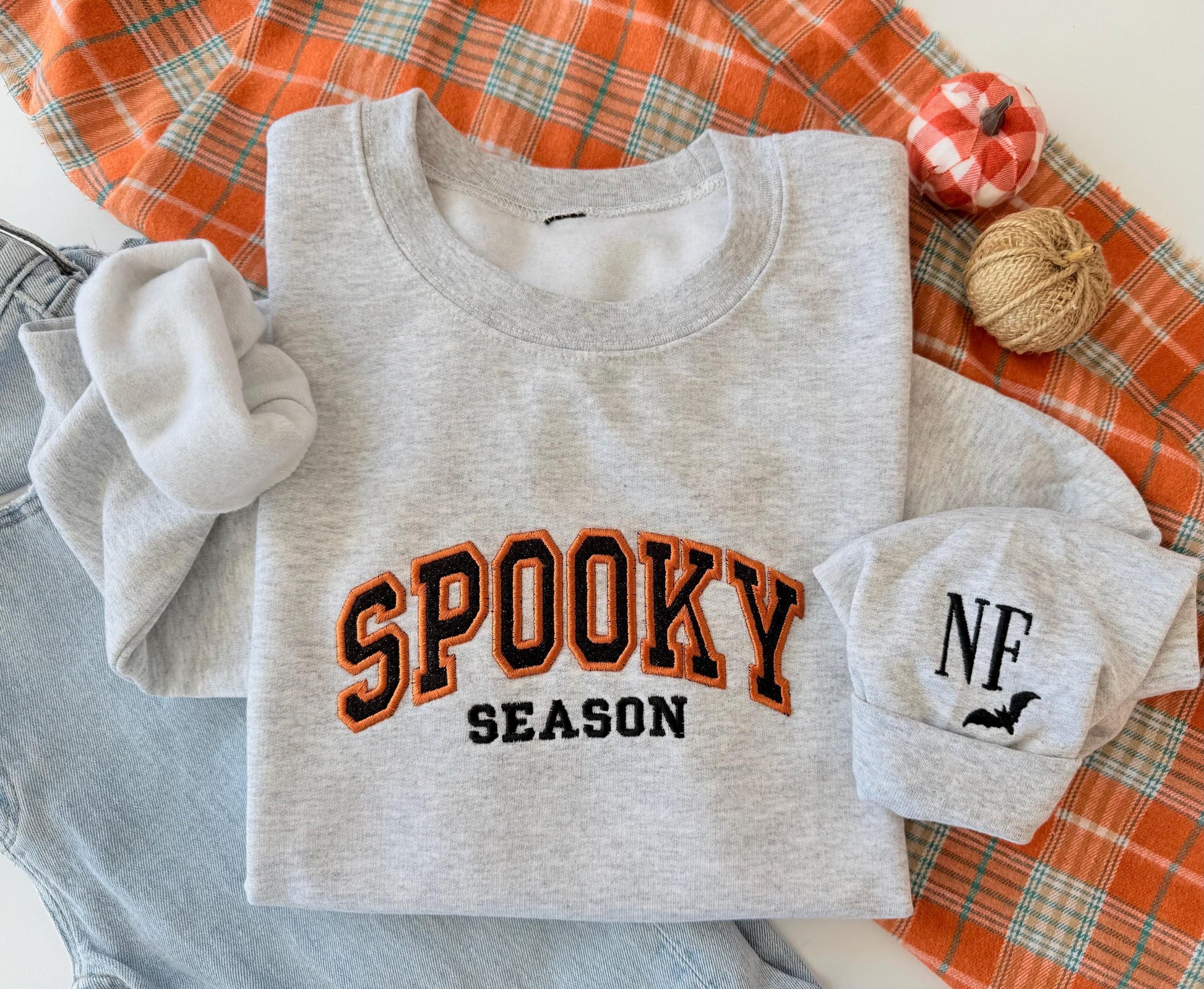 A light gray sweatshirt with "SPOOKY SEASON" embroidered in orange and black, and a cuff with "NF" and a bat, on an orange plaid blanket next to jeans and small decorative pumpkins.
