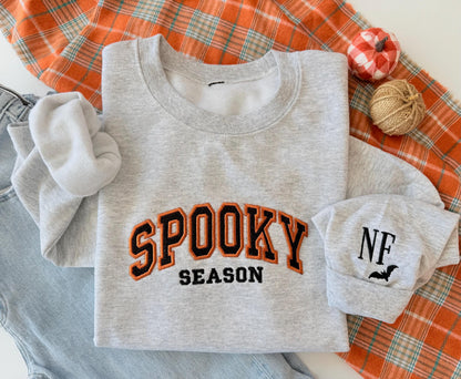 A light gray sweatshirt with "SPOOKY SEASON" embroidered in orange and black, and a cuff with "NF" and a bat, on an orange plaid blanket next to jeans and small decorative pumpkins.