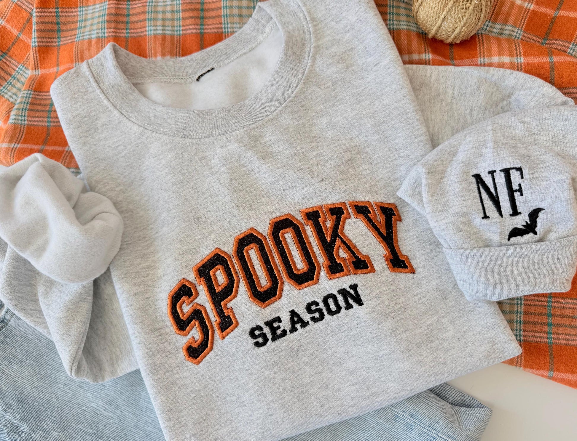 A light gray sweatshirt with "SPOOKY SEASON" embroidered in orange and black, and a cuff with "NF" and a bat, on an orange plaid blanket next to jeans and small decorative pumpkins.