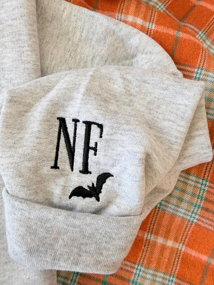 A close-up of a light gray sweatshirt cuff with embroidered black initials "NF" and a black bat icon, on an orange plaid blanket.