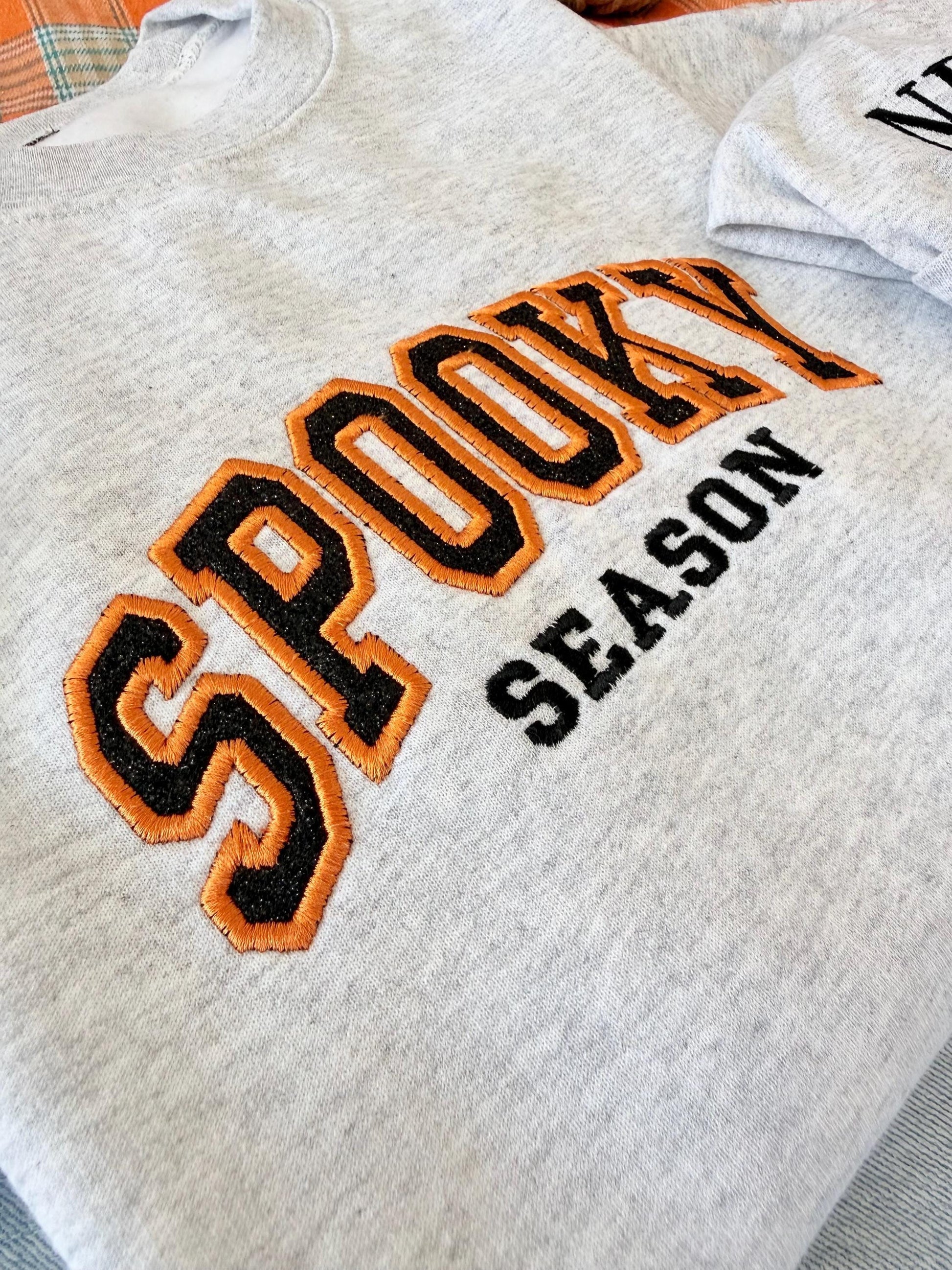 A close-up of a light gray sweatshirt with "SPOOKY SEASON" embroidered in orange and black, showing the texture of the embroidery.