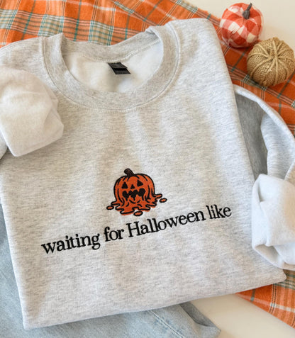 A light gray sweatshirt with an embroidered melting jack-o'-lantern and the text "waiting for Halloween like" is displayed on an orange plaid fabric next to two small decorative pumpkins.