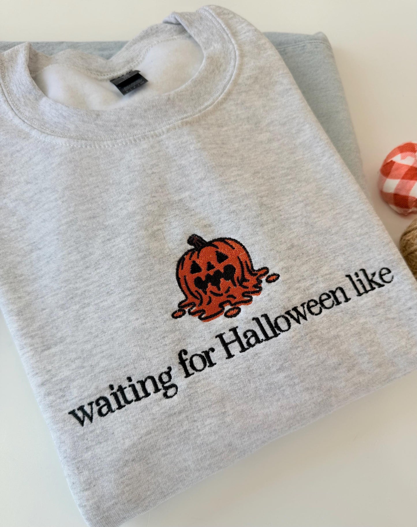 A light gray sweatshirt with an embroidered melting jack-o'-lantern and the text "waiting for Halloween like" is displayed on a light gray surface next to a small red and white plaid pumpkin.