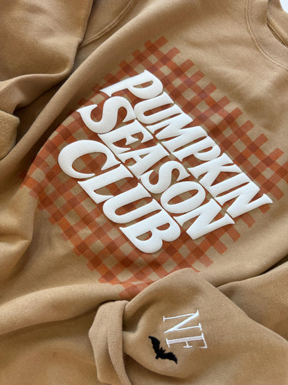 A close-up of a tan sweatshirt with "PUMPKIN SEASON CLUB" in white text over an orange gingham pattern, and "NF" with a black bat embroidered on the left sleeve.