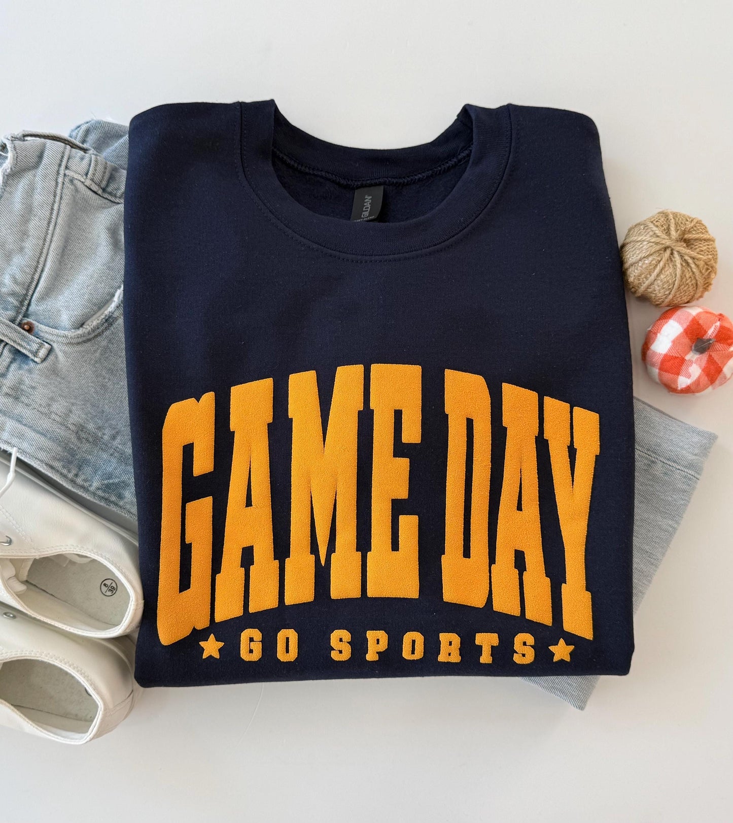 A navy blue crewneck sweatshirt with "GAME DAY" and "GO SPORTS" in orange embossed lettering, folded on a white surface next to light-wash jeans, white sneakers, and two small decorative gourds.