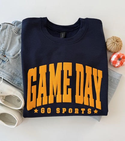 A navy blue crewneck sweatshirt with "GAME DAY" and "GO SPORTS" in orange embossed lettering, folded on a white surface next to light-wash jeans, white sneakers, and two small decorative gourds.