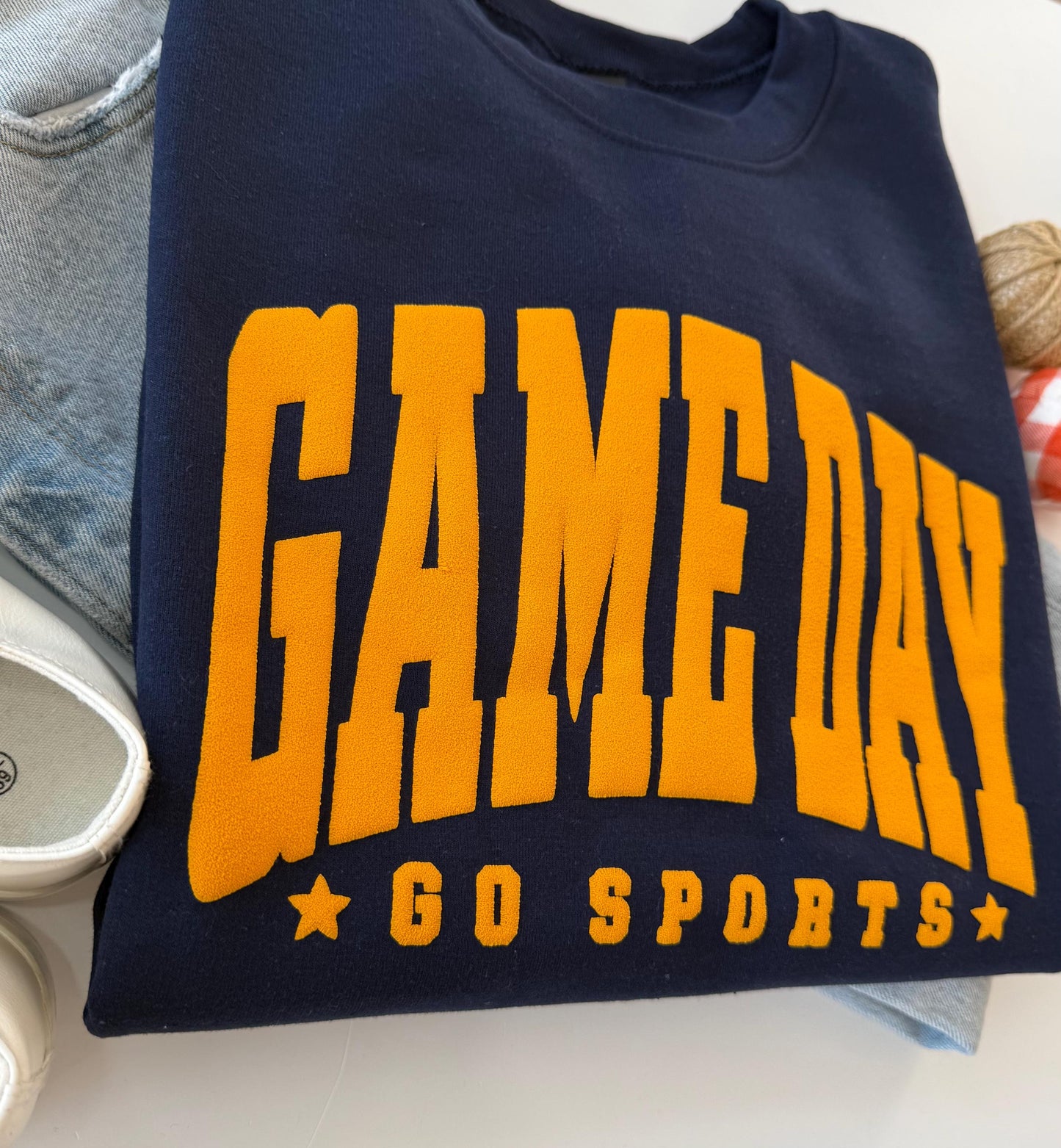 A close-up of a navy blue crewneck sweatshirt with "GAME DAY" and "GO SPORTS" in orange embossed lettering, folded on a white surface next to light-wash jeans and a red and white checkered gourd.