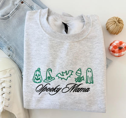 A light gray crewneck sweatshirt with green embroidered Halloween icons and "Spooky Mama" in black script, folded on a white surface with jeans and decorative pumpkins.