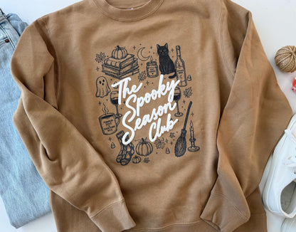 A tan crewneck sweatshirt with "The Spooky Season Club" in white script, surrounded by black line drawings of Halloween and fall items, including a black cat, pumpkins, and a broom, laid flat next to blue jeans and a white shoe.