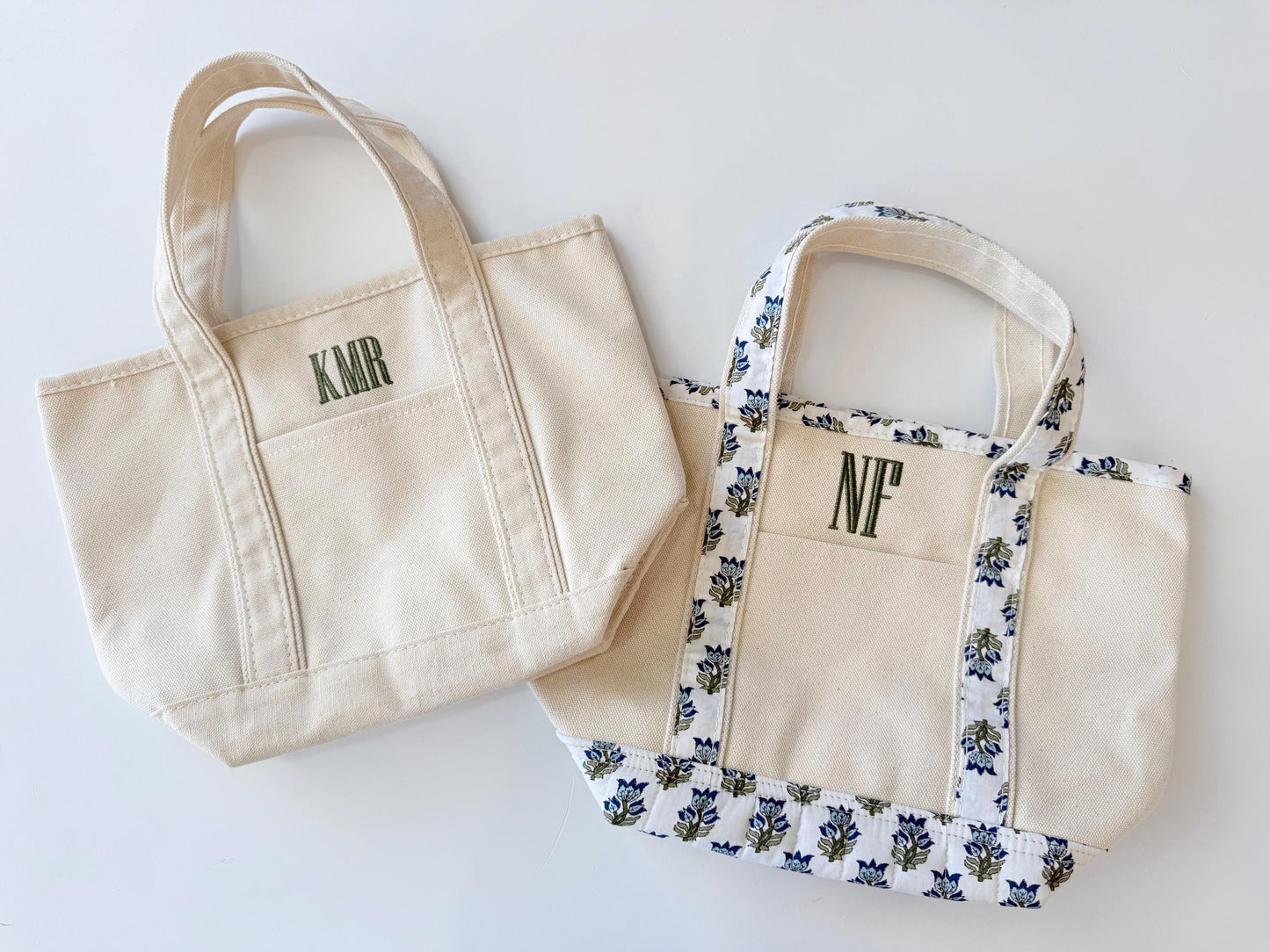 Two small canvas tote bags are shown on a white surface, one plain with "KMR" in green, and one with a blue floral pattern and "NF" in green.