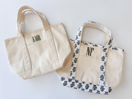 Two small canvas tote bags are shown on a white surface, one plain with "KMR" in green, and one with a blue floral pattern and "NF" in green.
