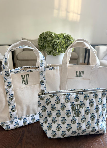 Three canvas items are on a dark wood table: a small tote with blue floral trim and "NF", a plain tote with "KMR", and a quilted pouch with blue floral pattern and "NF".