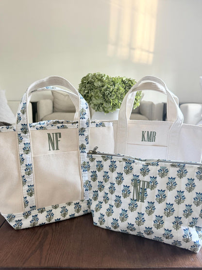 Three canvas items are on a dark wood table: a small tote with blue floral trim and "NF", a plain tote with "KMR", and a quilted pouch with blue floral pattern and "NF".