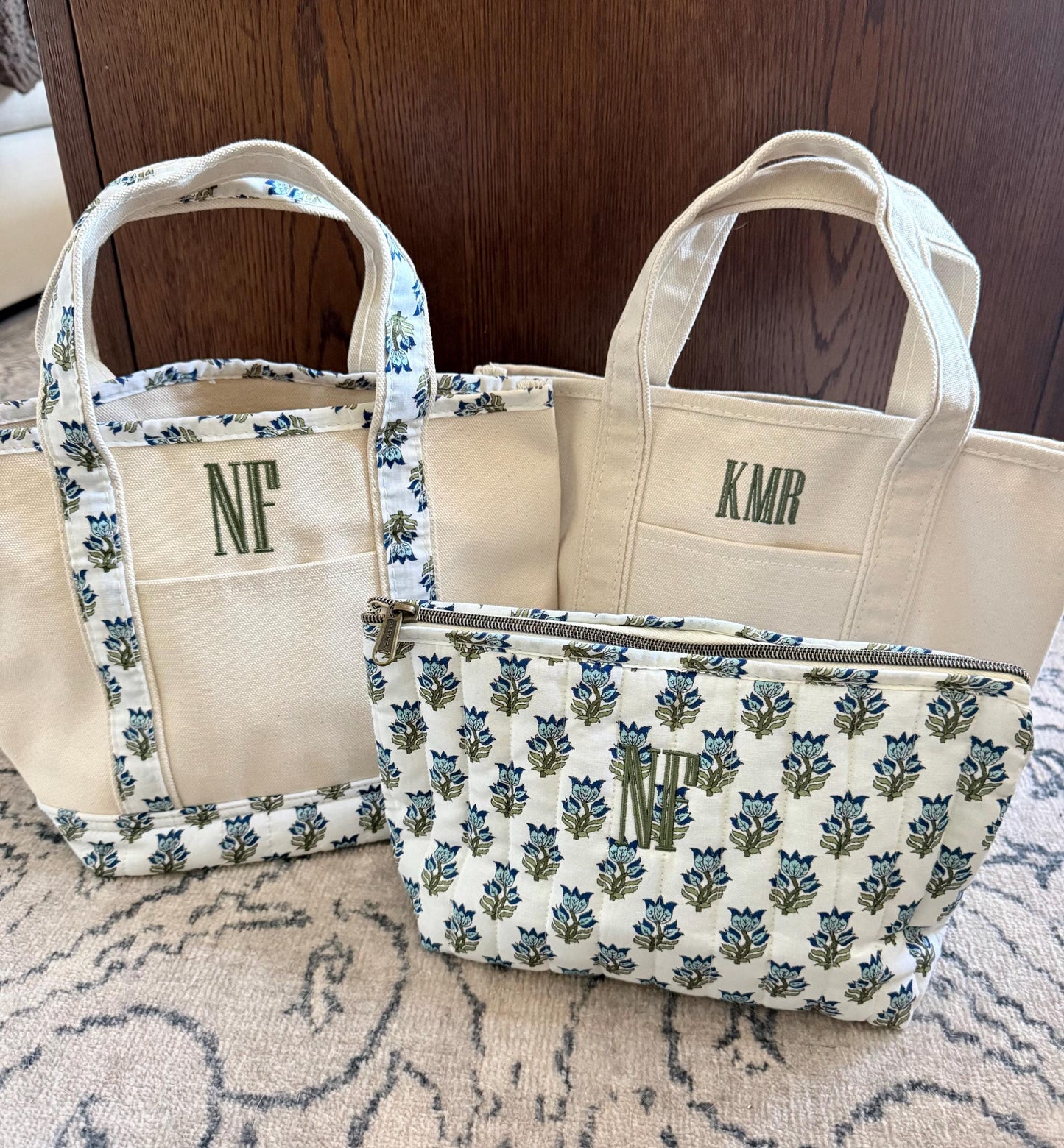 Three canvas items are on a light rug in front of a dark wood surface: a small tote with blue floral trim and "NF", a plain tote with "KMR", and a quilted pouch with blue floral pattern and "NF".