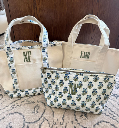 Three canvas items are on a light rug in front of a dark wood surface: a small tote with blue floral trim and "NF", a plain tote with "KMR", and a quilted pouch with blue floral pattern and "NF".