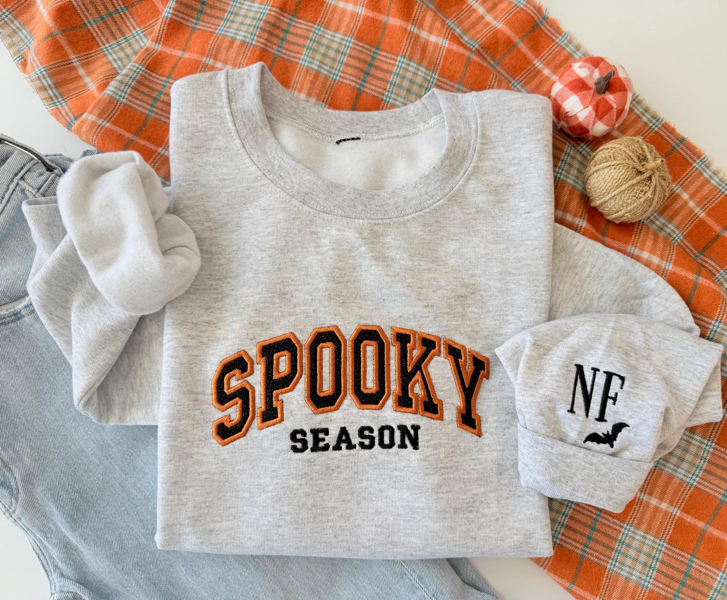 A light gray sweatshirt with "SPOOKY SEASON" embroidered in orange and black, a cuff with "NF" and a bat, on an orange plaid blanket next to jeans and small decorative pumpkins.