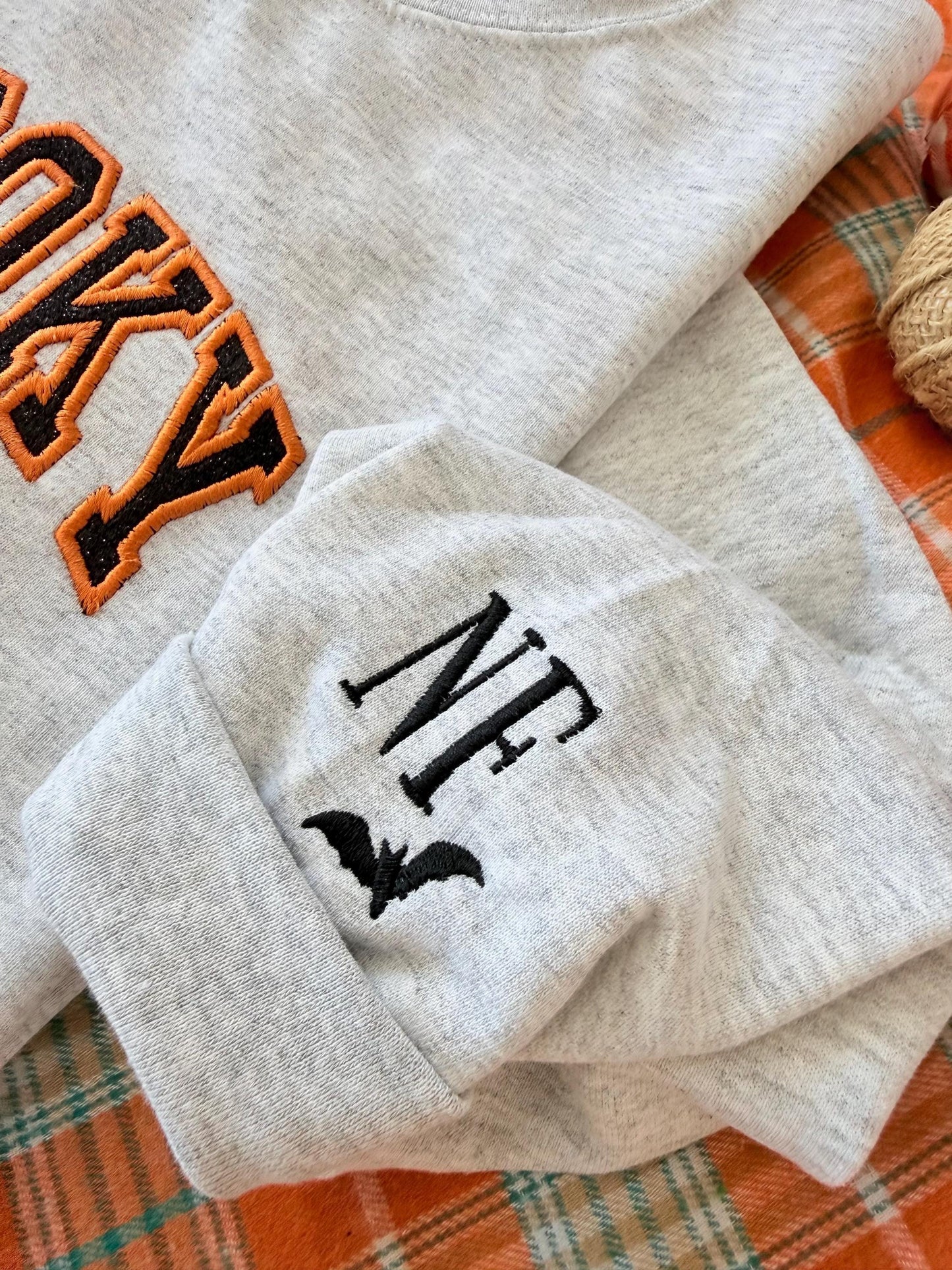 A close-up of a light gray sweatshirt cuff with embroidered black initials "NF" and a black bat icon, next to the orange and black "SPOOKY" embroidery.