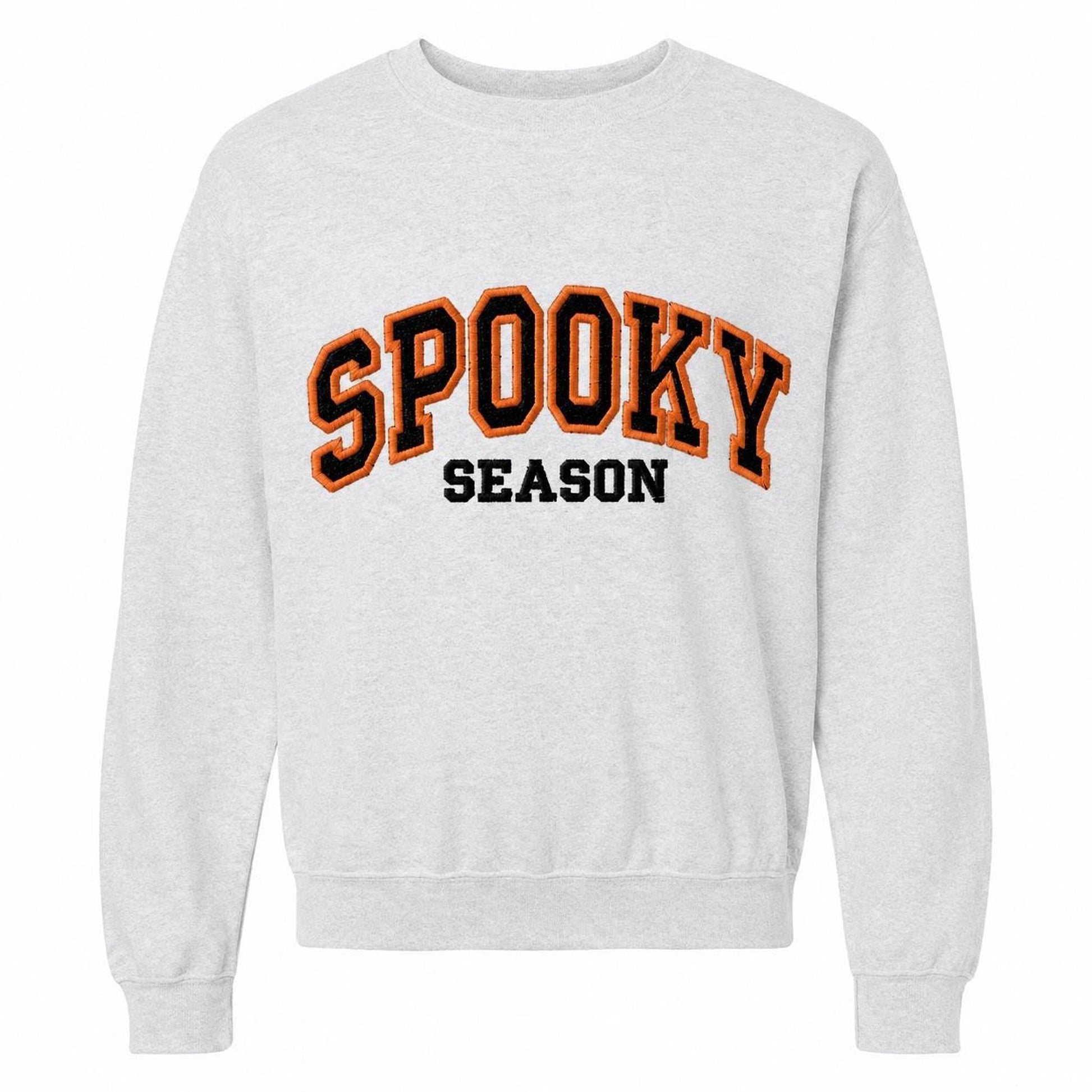 A light gray crewneck sweatshirt with "SPOOKY SEASON" embroidered across the chest in orange and black varsity-style letters.