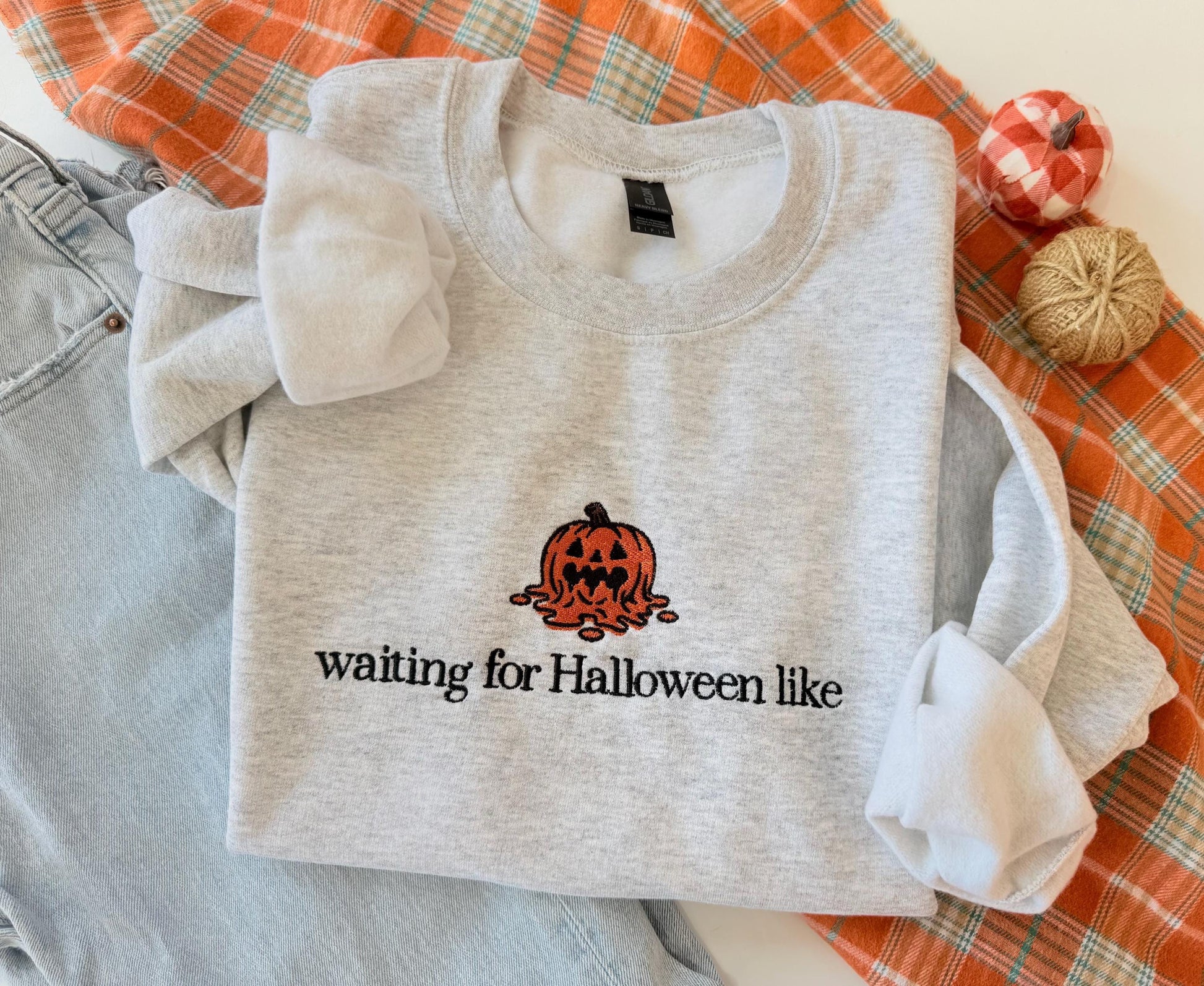 A light gray sweatshirt with an embroidered melting jack-o'-lantern and the text "waiting for Halloween like" is displayed on an orange plaid fabric next to light wash jeans and two small decorative pumpkins.
